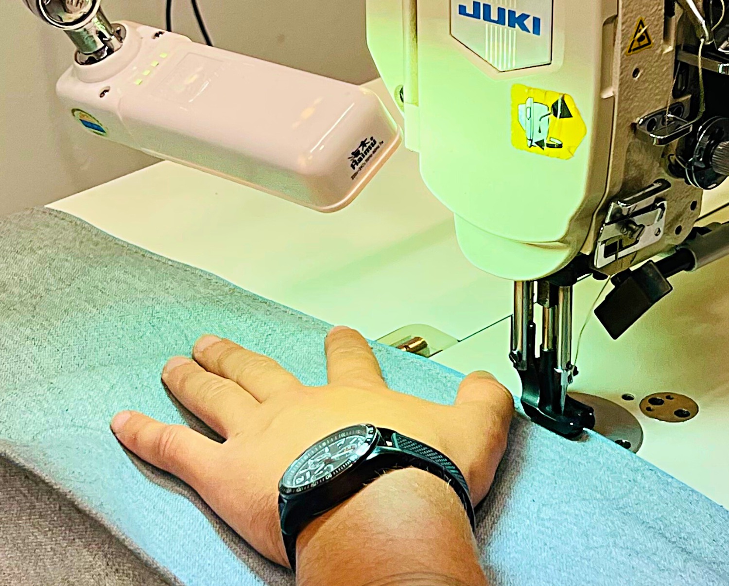 Szara tkanina przeszywana na maszynie JUKI, dłoń z czarnym zegarkiem przytrzymuje materiał pod igłą.