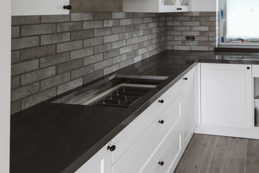 Ciemny blat kuchenny z wycięciem na zlew, białe szafki z czarnymi gałkami, ściana wyłożona szarą cegłą, widoczne gniazdka elektryczne.