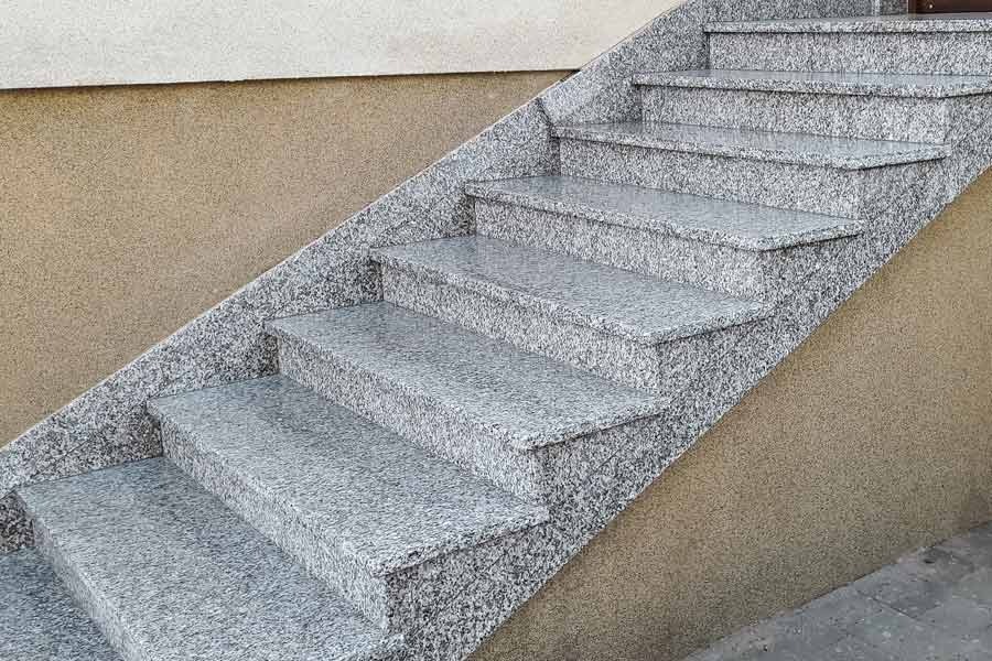 Zewnętrzne schody z szarego granitu prowadzące w górę, wykończone takim samym materiałem, przylegające do ściany o beżowej, chropowatej fakturze.
