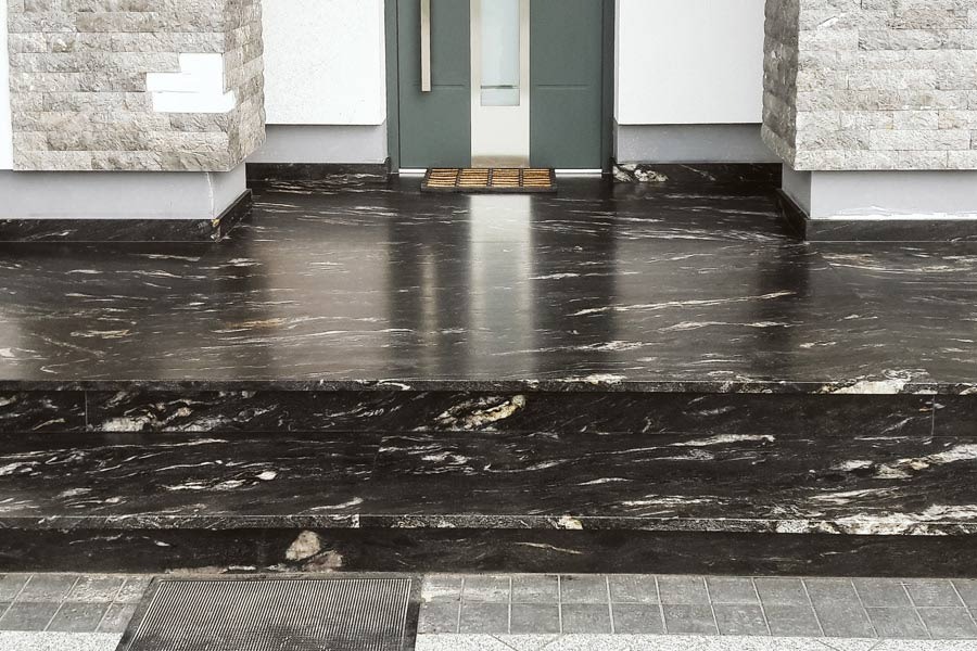 Polerowane, czarne marmurowe schody wejściowe z białymi żyłami, prowadzące do szarych drzwi wejściowych z wycieraczką, obok kolumny z kamienia elewacyjnego.