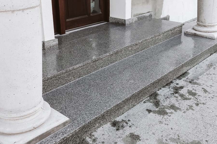 Dwa stopnie wejściowe z szarego lastryko, prowadzące do ciemnych drzwi, z kolumnami po bokach, widoczne ślady wilgoci na powierzchni.