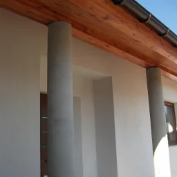 Fragment domu z elewacją z jasnego tynku strukturalnego, drewnianą podbitką dachu i okrągłymi, betonowymi słupami przy wejściu.