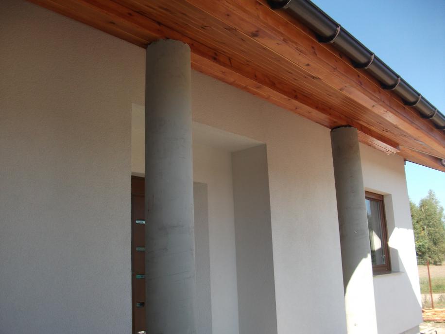 Fragment domu z elewacją z jasnego tynku strukturalnego, drewnianą podbitką dachu i okrągłymi, betonowymi słupami przy wejściu.