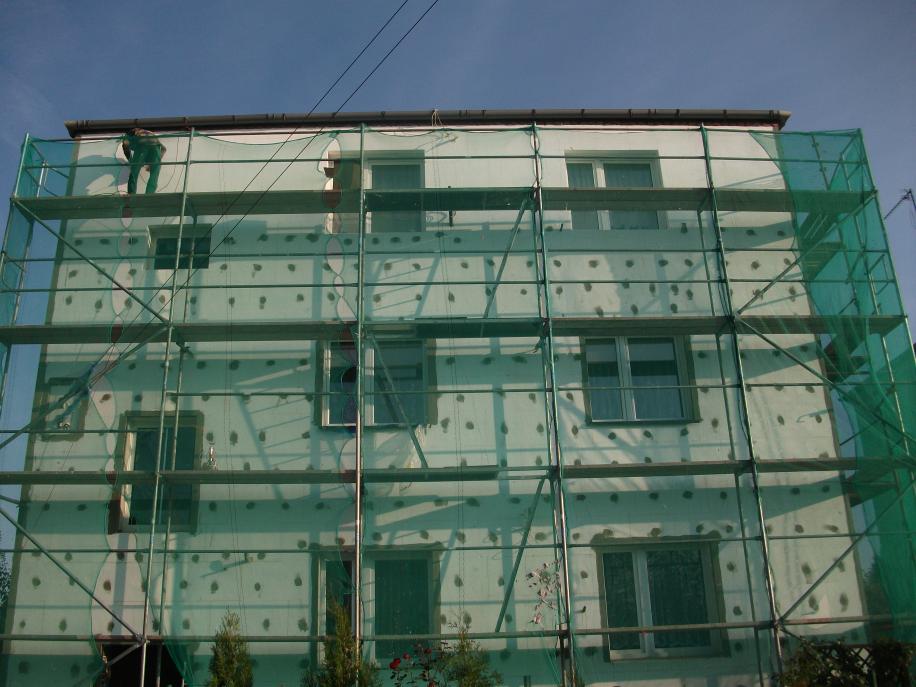 Docieplanie elewacji budynku styropianem w Zbietce, widoczne rusztowania i siatka ochronna, pracownik na górnym poziomie.