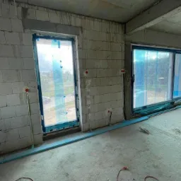 Surowy stan wnętrza budynku w trakcie prac: widoczne okna zabezpieczone folią, ściany z bloczków, wystające kable elektryczne i puszki pod gniazdka, nie wykończona podłoga.