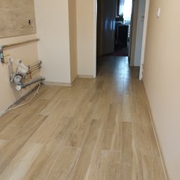 Remwik - Wyłożona płytkami imitującymi drewno podłoga w korytarzu, widoczne rury instalacji wodnej przy ścianie oraz fragment ściany z przygotowanym otworem pod gniazdko.