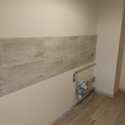 Remwik - Fragment ściany w trakcie remontu kuchni, z ułożonym pasem płytek imitujących drewno, widoczne rury instalacji wodnej i przygotowane punkty elektryczne.