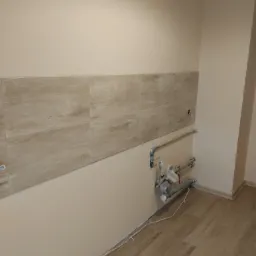 Fragment ściany w trakcie remontu kuchni, z ułożonym pasem płytek imitujących drewno, widoczne rury instalacji wodnej i przygotowane punkty elektryczne.