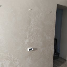 Remwik - Ściana w odcieniach beżu z dekoracyjną strukturą, widoczne puszki elektryczne i haki, fragment podłogi z paneli.