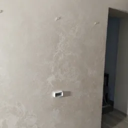 Ściana w odcieniach beżu z dekoracyjną strukturą, widoczne puszki elektryczne i haki, fragment podłogi z paneli.