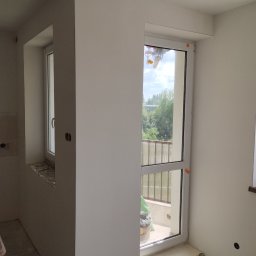 Remwik - Świeżo wyszpachlowane ściany w pomieszczeniu z oknem i drzwiami balkonowymi, widoczne niedoskonałości podłoża przed malowaniem.