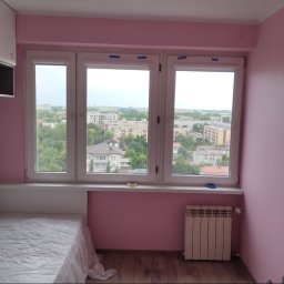 Remwik - Pokój po odświeżeniu: Różowe ściany, nowe okno z widokiem na miasto, zabezpieczone taśmą malarską, zakryte meble, grzejnik, drewniana podłoga.