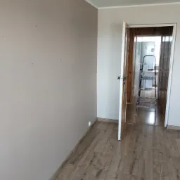 Świeżo pomalowany pokój z beżowymi ścianami i drewnianą podłogą, widoczne ślady po szpachlowaniu, drabina w przejściu do kuchni.