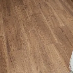 Ułożone panele podłogowe imitujące drewno, z widocznymi listwami przypodłogowymi. Fragment ściany z szarą, surową powierzchnią nad listwą.