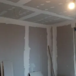 Pokój w trakcie prac wykończeniowych: ściany pokryte płytami gipsowo-kartonowymi, widoczne łączenia szpachlowane, poziomica przy ścianie, sufit również z płyt g-k, okno zabezpieczone taśmą malarską.