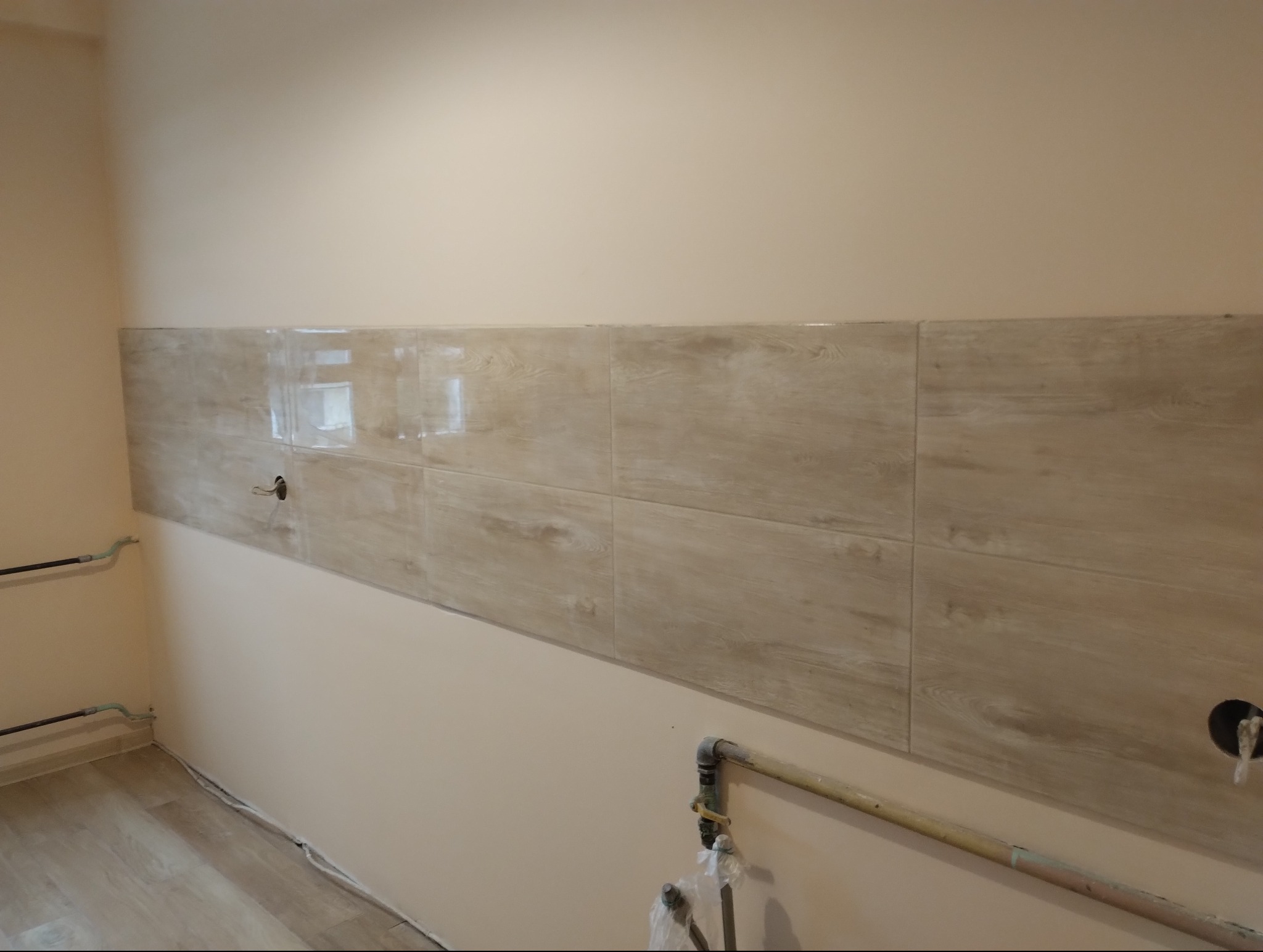 Fragment ściany w kuchni z ułożonymi płytkami imitującymi drewno, widoczne instalacje hydrauliczne i elektryczne przed wykończeniem.