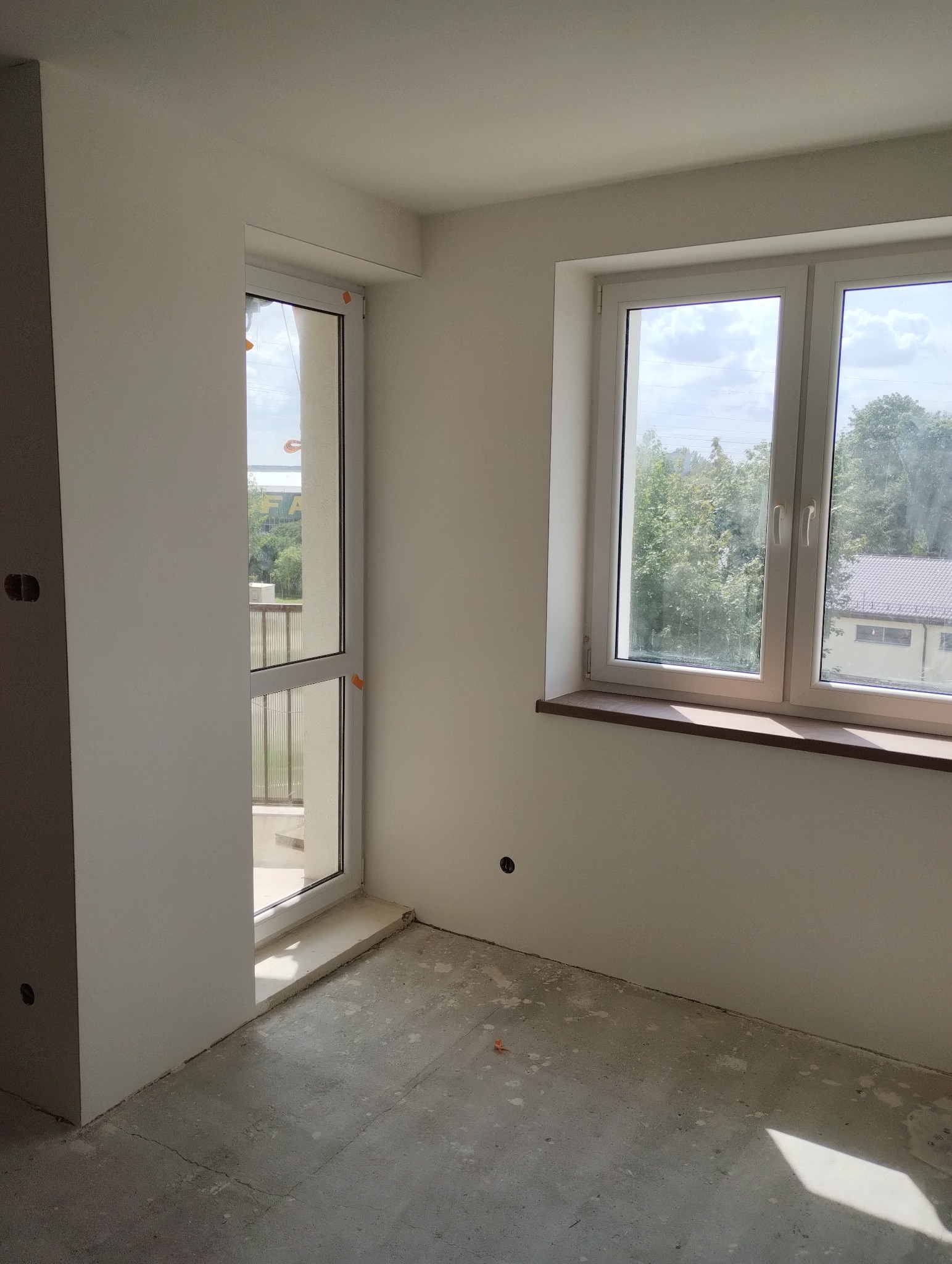 Wnętrze pokoju w trakcie prac wykończeniowych, widoczne gładkie, białe ściany, okno z widokiem na zieleń oraz surowa betonowa podłoga z drobnymi zabrudzeniami.