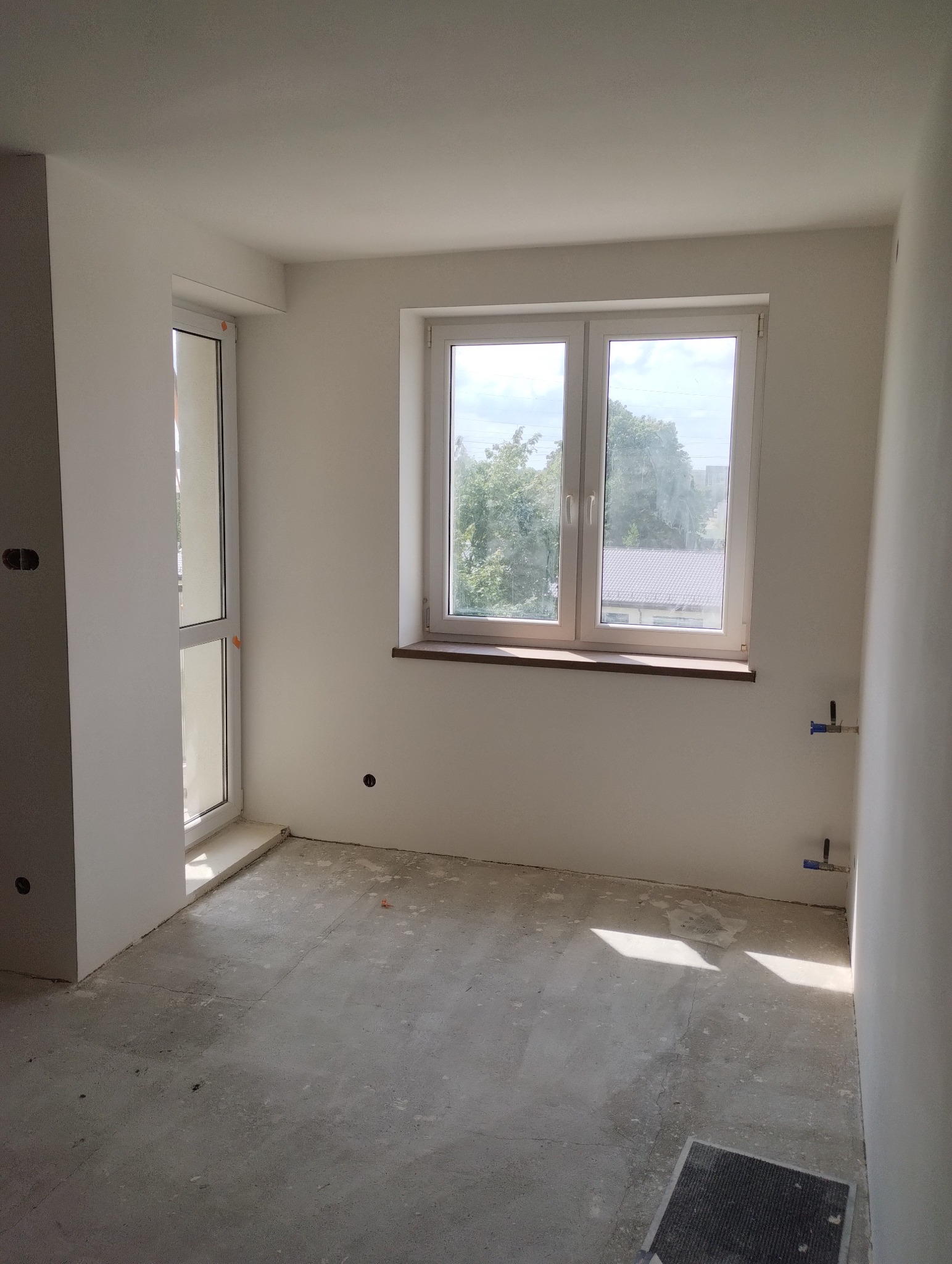 Surowe wnętrze pokoju z oknem i drzwiami balkonowymi, ściany pokryte świeżą, białą farbą, widoczne przyłącza wodne, podłoga betonowa przed wylewką.