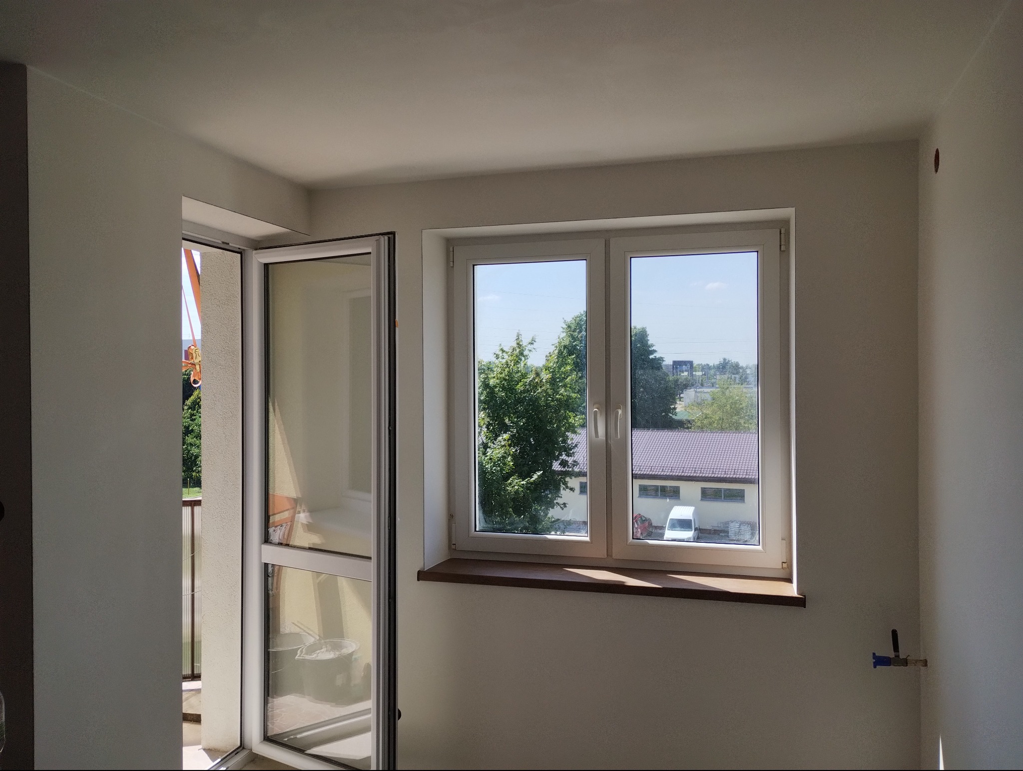 Wnętrze pokoju po remoncie z białymi ścianami, oknem z widokiem na zewnątrz i otwartymi drzwiami balkonowymi, widoczne wiaderka z farbą.