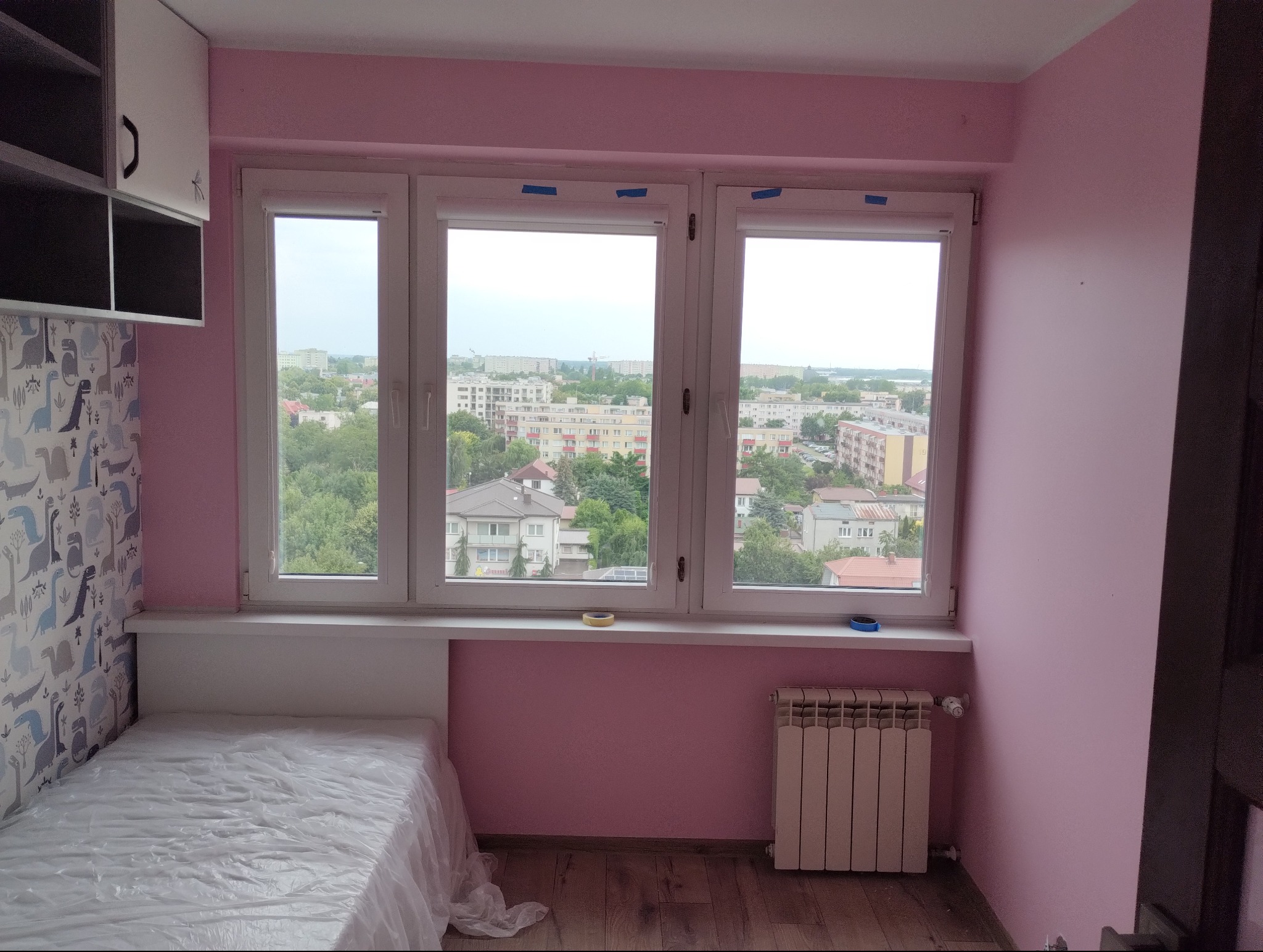 Pokój po odświeżeniu: Różowe ściany, nowe okno z widokiem na miasto, zabezpieczone taśmą malarską, zakryte meble, grzejnik, drewniana podłoga.