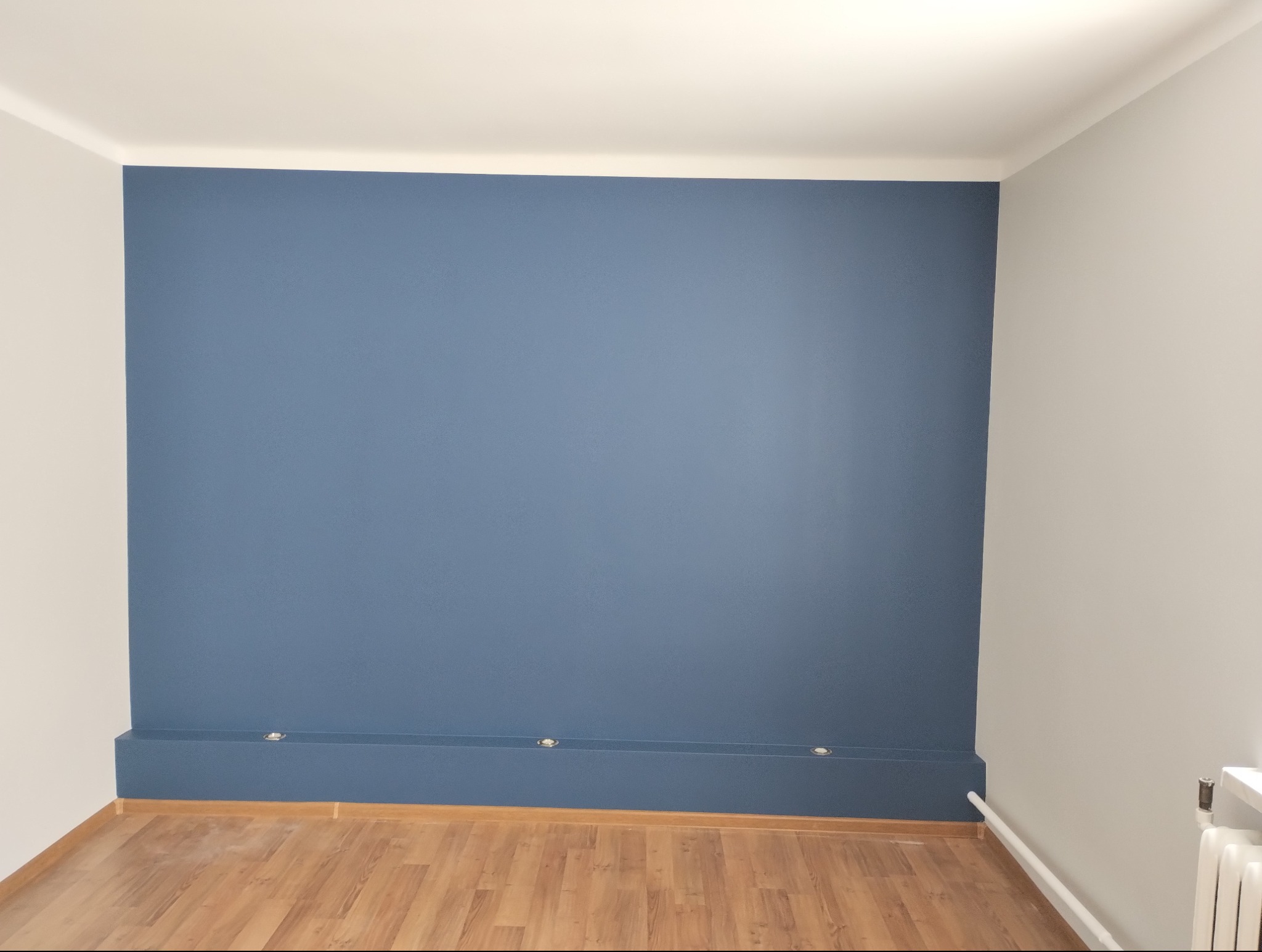 Świeżo pomalowana na niebiesko ściana z listwą przypodłogową z wbudowanymi punktowymi lampkami, widoczny fragment parkietu i pomalowana na szaro sąsiednia ściana.