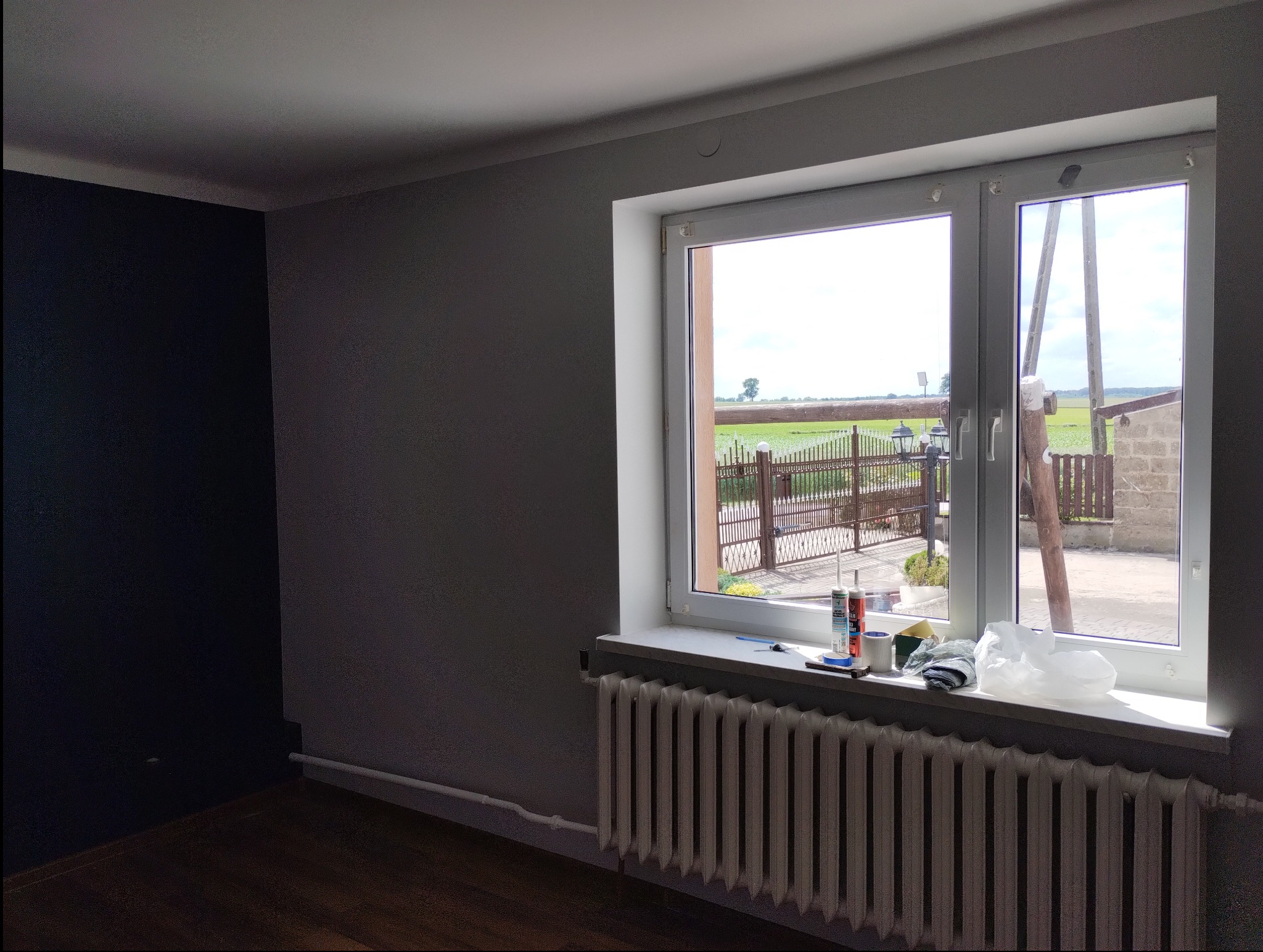 Pokój po malowaniu: jedna ściana w kolorze grafitowym, reszta szara, w oknie widok na zewnątrz, na parapecie akcesoria malarskie.