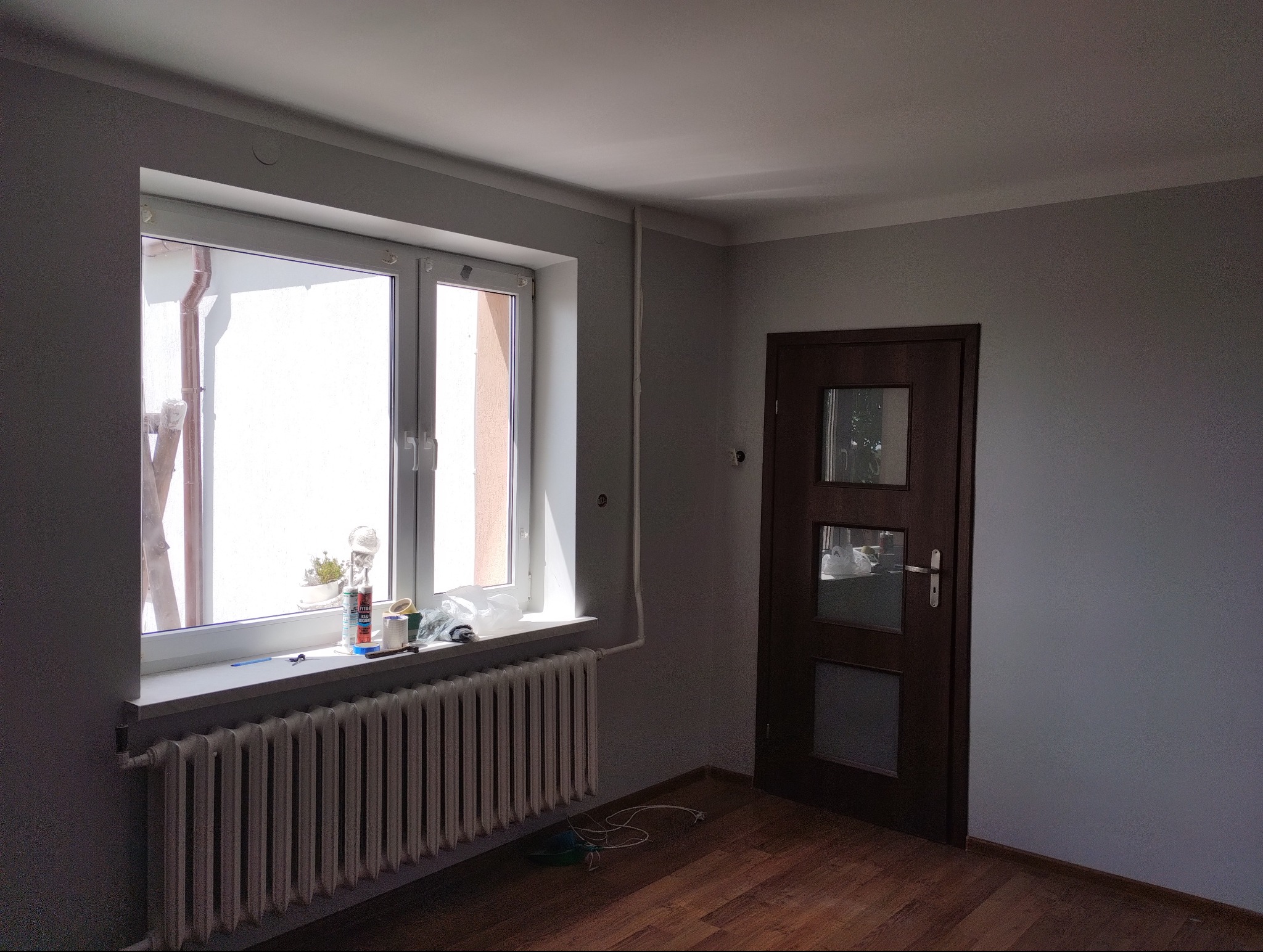 Pokój po odświeżeniu ścian w szarym odcieniu, widoczny grzejnik pod oknem z akcesoriami malarskimi na parapecie i drewniana podłoga.