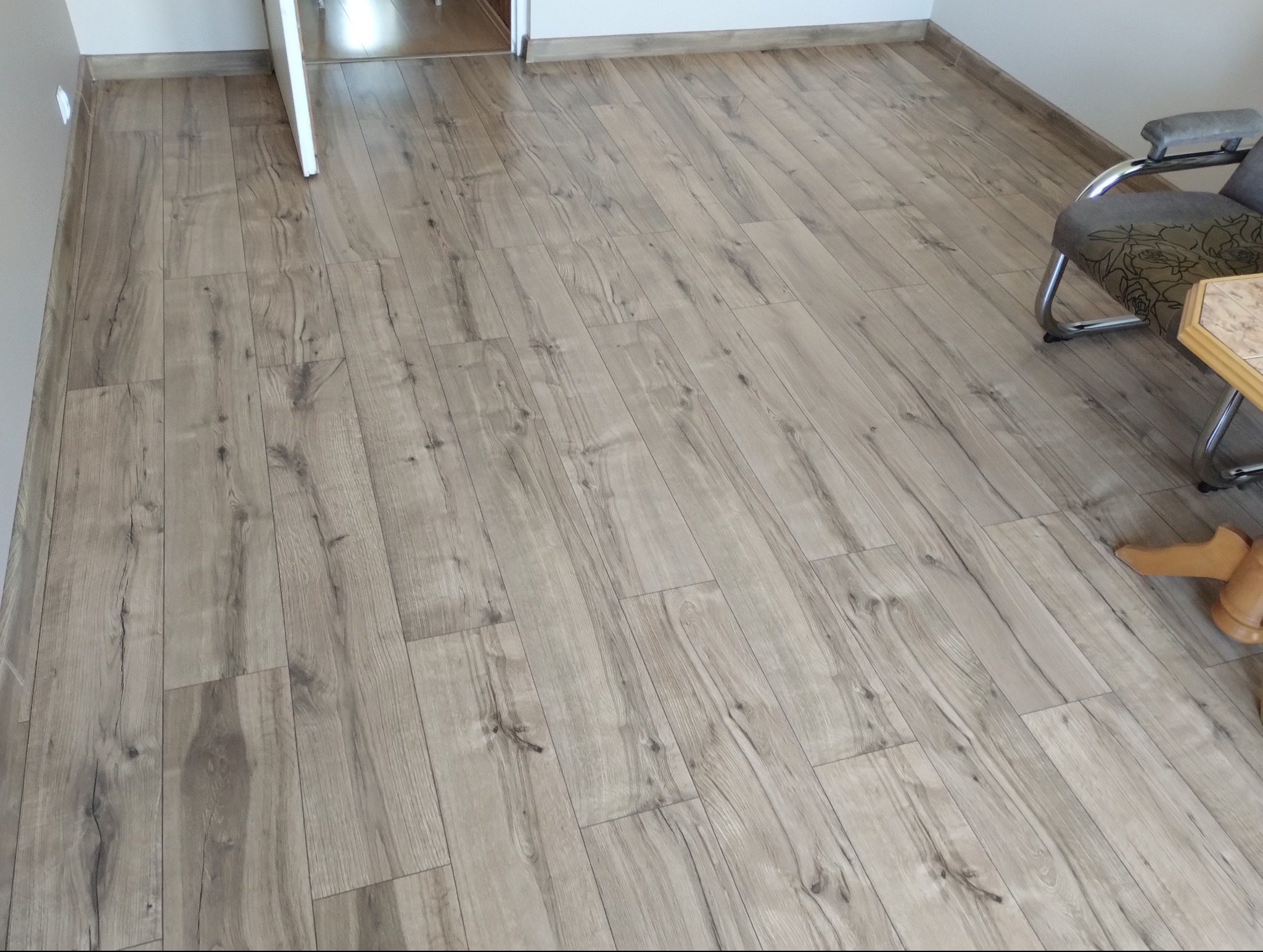 Ułożona podłoga z paneli imitujących drewno w odcieniach szarości i beżu, widok z góry na całą powierzchnię z fragmentem krzesła i stołu w rogu pomieszczenia.