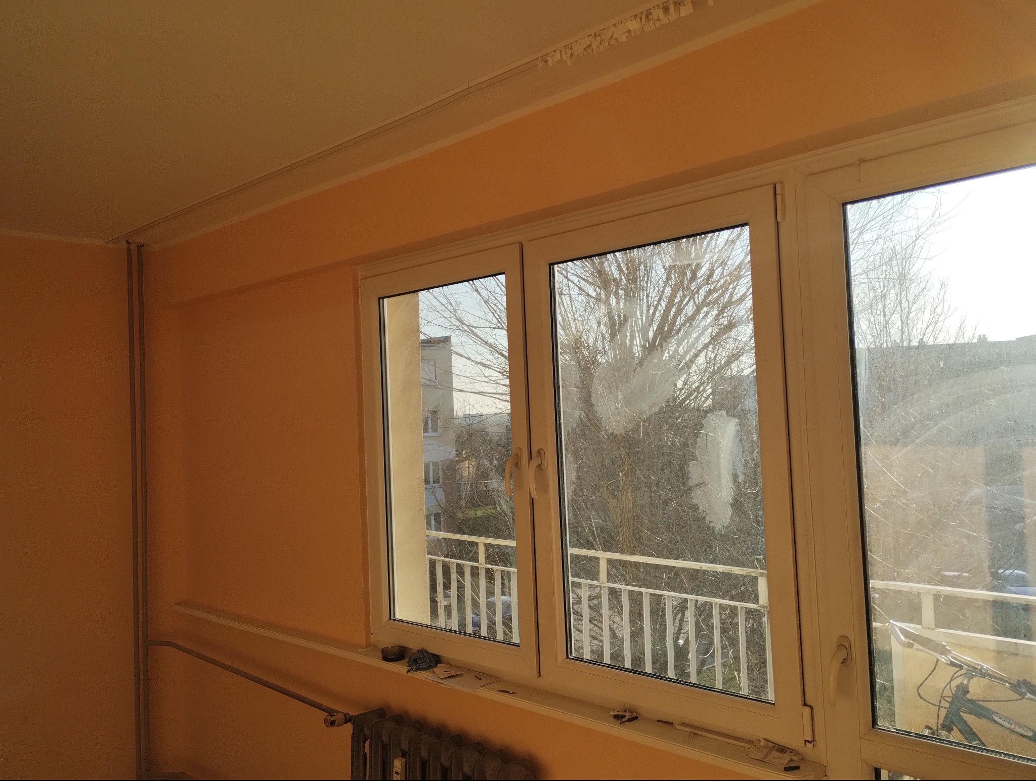 Pokój z pomarańczowymi ścianami przed malowaniem, widoczne okno z zabrudzeniami, grzejnik i listwy przypodłogowe.