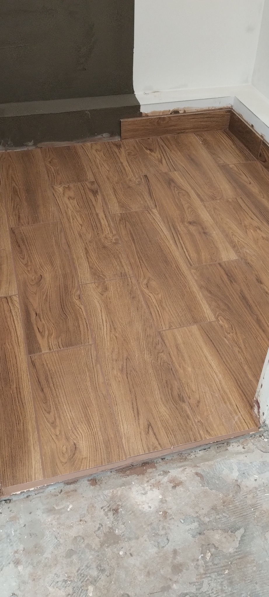 Ułożone panele podłogowe imitujące drewno, z widocznymi listwami przypodłogowymi. Fragment ściany z szarą, surową powierzchnią nad listwą.