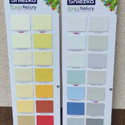 Dwa ekspozytory z paletą barw farb lateksowych Śnieżka Barwy Natury, prezentujące odcienie od bieli i beżu po niebieskie i szarości, umieszczone na tle ściany i kamienistego podłoża.