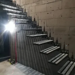 Montaż metalowych schodów wspornikowych z elementami dekoracyjnymi w surowym wnętrzu, widoczny laserowy niwelator i surowa ściana z betonu komórkowego.