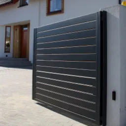 Nowoczesna, przesuwna brama wjazdowa z poziomymi panelami w kolorze antracytowym, zamontowana przy jasnym, dwupiętrowym domu z drewnianą stolarką okienną i brukowanym podjazdem.