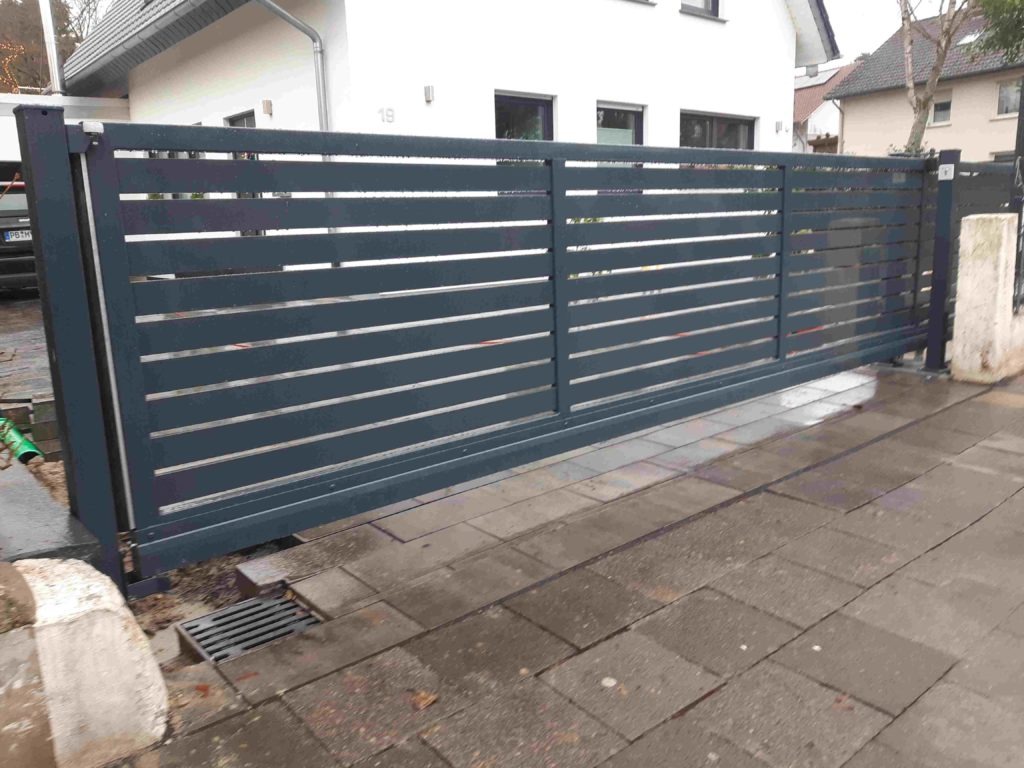 Nowoczesna, przesuwna brama wjazdowa z poziomych, metalowych profili, szara, widok z zewnątrz, na pierwszym planie kostka brukowa i kratka ściekowa.