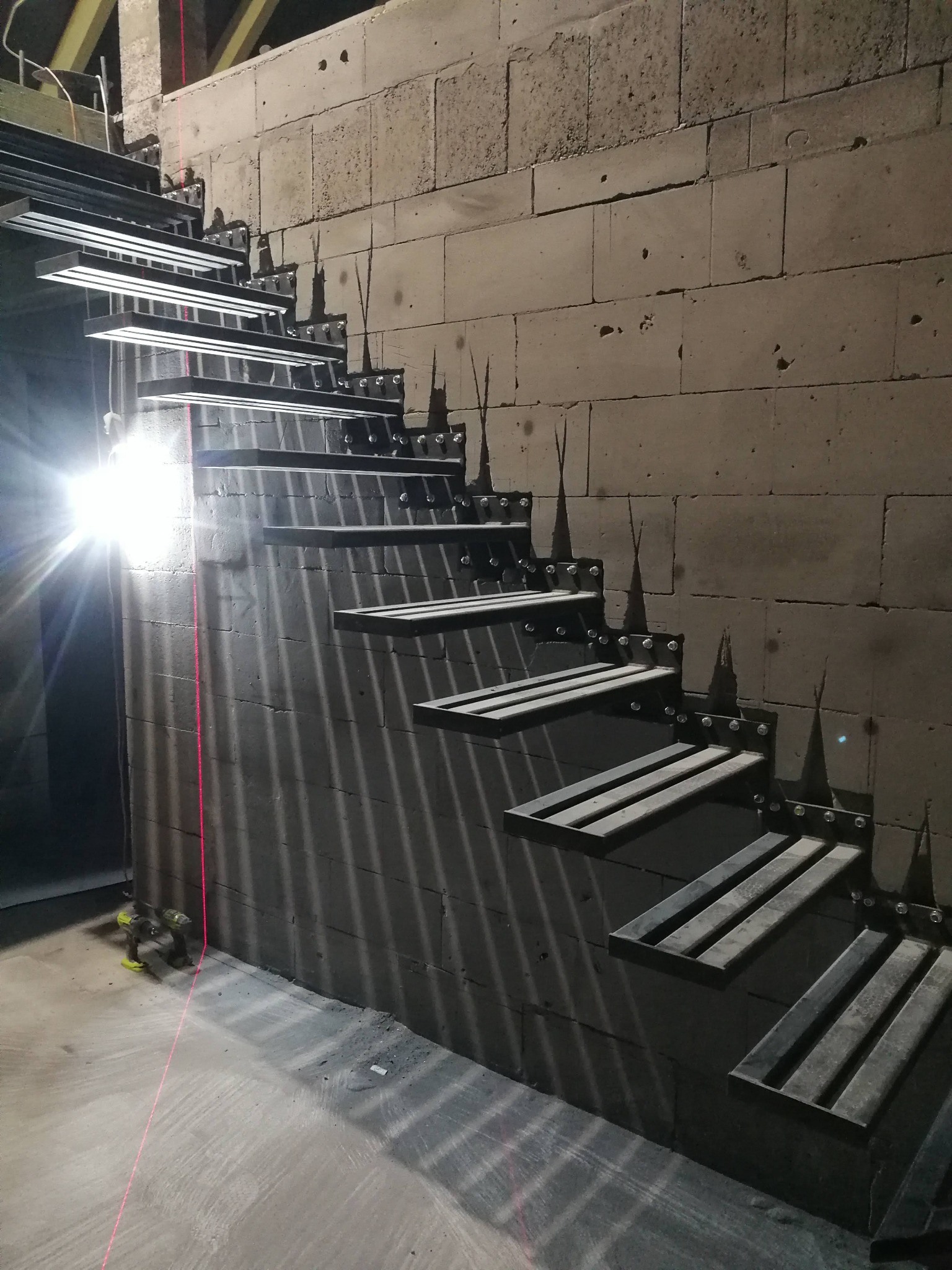 Montaż metalowych schodów wspornikowych z elementami dekoracyjnymi w surowym wnętrzu, widoczny laserowy niwelator i surowa ściana z betonu komórkowego.