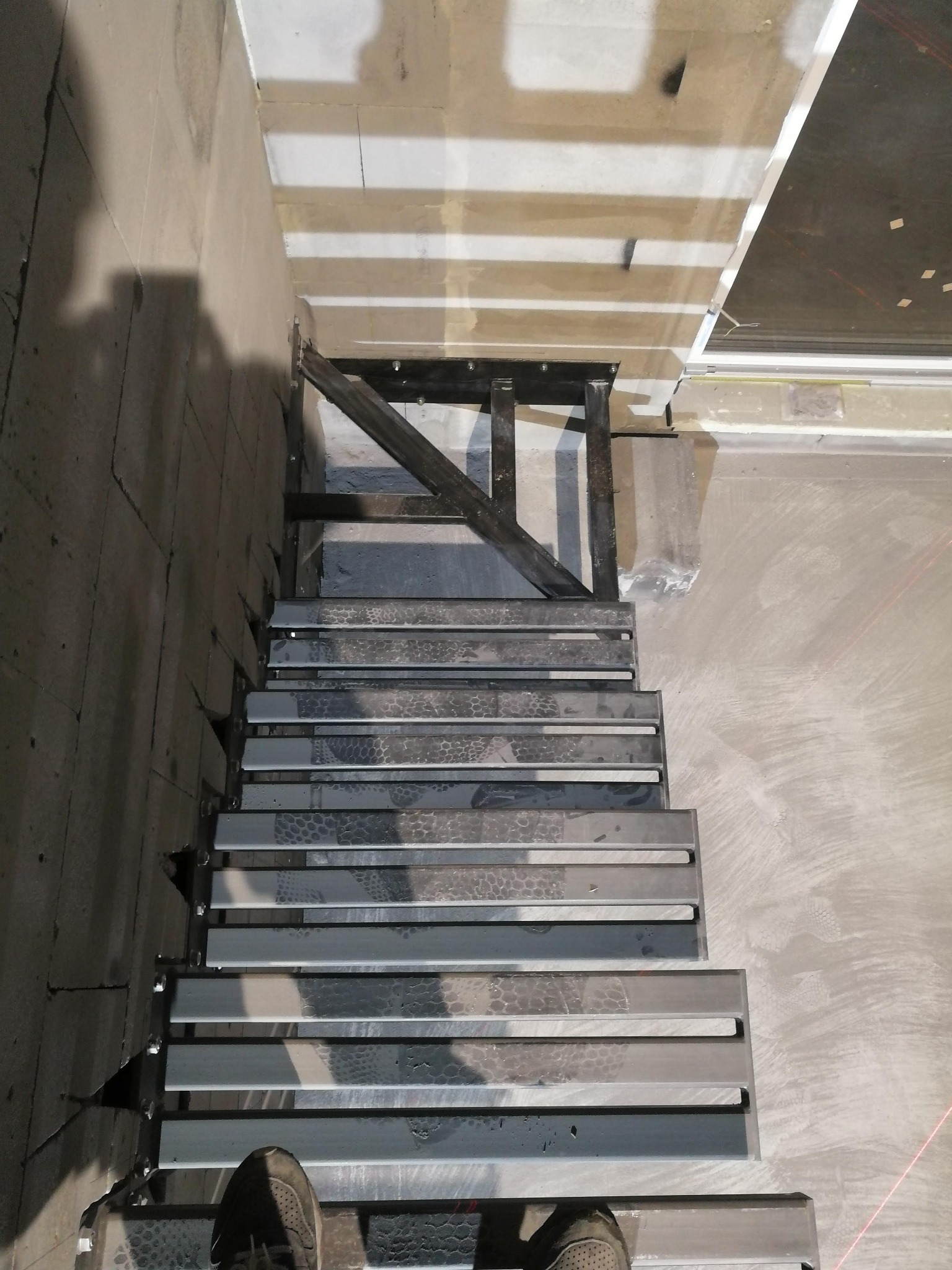 Surowe, metalowe schody z widocznymi spawami i elementami mocującymi, w trakcie montażu w surowym wnętrzu. Widoczne ślady po spawaniu i niedoskonałości powierzchni.