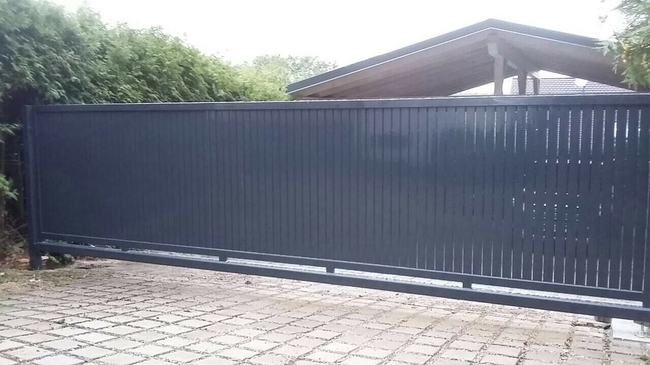 Przesuwna brama wjazdowa z pionowych metalowych profili w kolorze grafitowym, widoczna na tle kostki brukowej i zieleni, z zadaszeniem w tle.