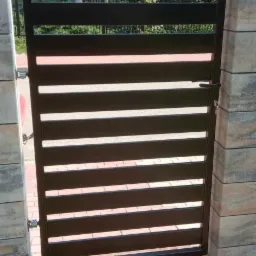 Nowoczesna, brązowa furtka z poziomych profili, zamontowana między kamiennymi słupkami. Widoczne detale zamka i zawiasów. Tło stanowi dom i zieleń.