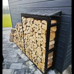 Metalowy regał na drewno opałowe z daszkiem, wypełniony pociętymi kawałkami drewna, stojący na szarym bruku przy ciemnej, drewnianej ścianie budynku.