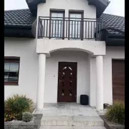 Dwukondygnacyjny dom z balkonem z czarną balustradą, kolumnami przy wejściu i ciemnobrązowymi drzwiami. Widoczne schody wejściowe i roślinność.