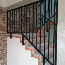 Czarna, metalowa balustrada schodowa z dekoracyjnymi elementami, zamontowana na zabezpieczonych schodach wewnątrz budynku. Widoczna ceglana ściana i jasne ściany.