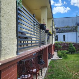 KM Patryk Szymański - Nowoczesna, pozioma balustrada balkonowa w kolorze antracytu, z widokiem na meble ogrodowe i zadbany trawnik. Elementy dekoracyjne: kwiaty w donicach.