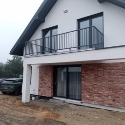 KM Patryk Szymański - Nowoczesny dom z balkonem i czarną balustradą. Elewacja z białego tynku i cegły klinkierowej. Widoczny fragment podjazdu i samochód. Ujęcie z dołu, pochmurny dzień.