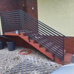 KM Patryk Szymański - Nowoczesna, pozioma balustrada schodowa z metalu w kolorze grafitowym, zamontowana przy wejściu do domu z ceglaną elewacją i beżowym tynkiem.