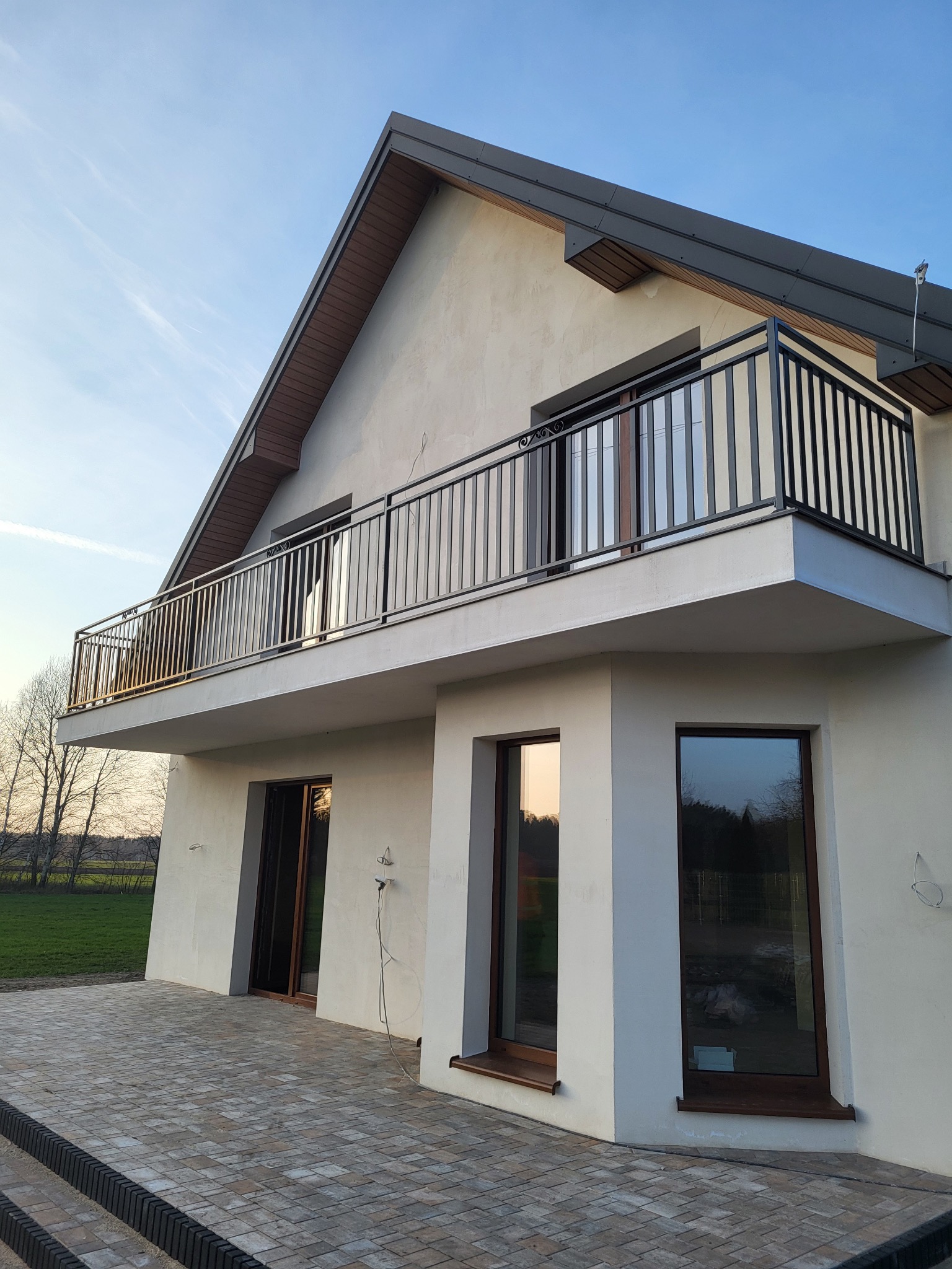 Nowoczesny dom z balkonem i czarną, metalową balustradą. Widoczne okna z ciemnymi ramami i jasna elewacja. Brukowany taras i trawnik w tle.