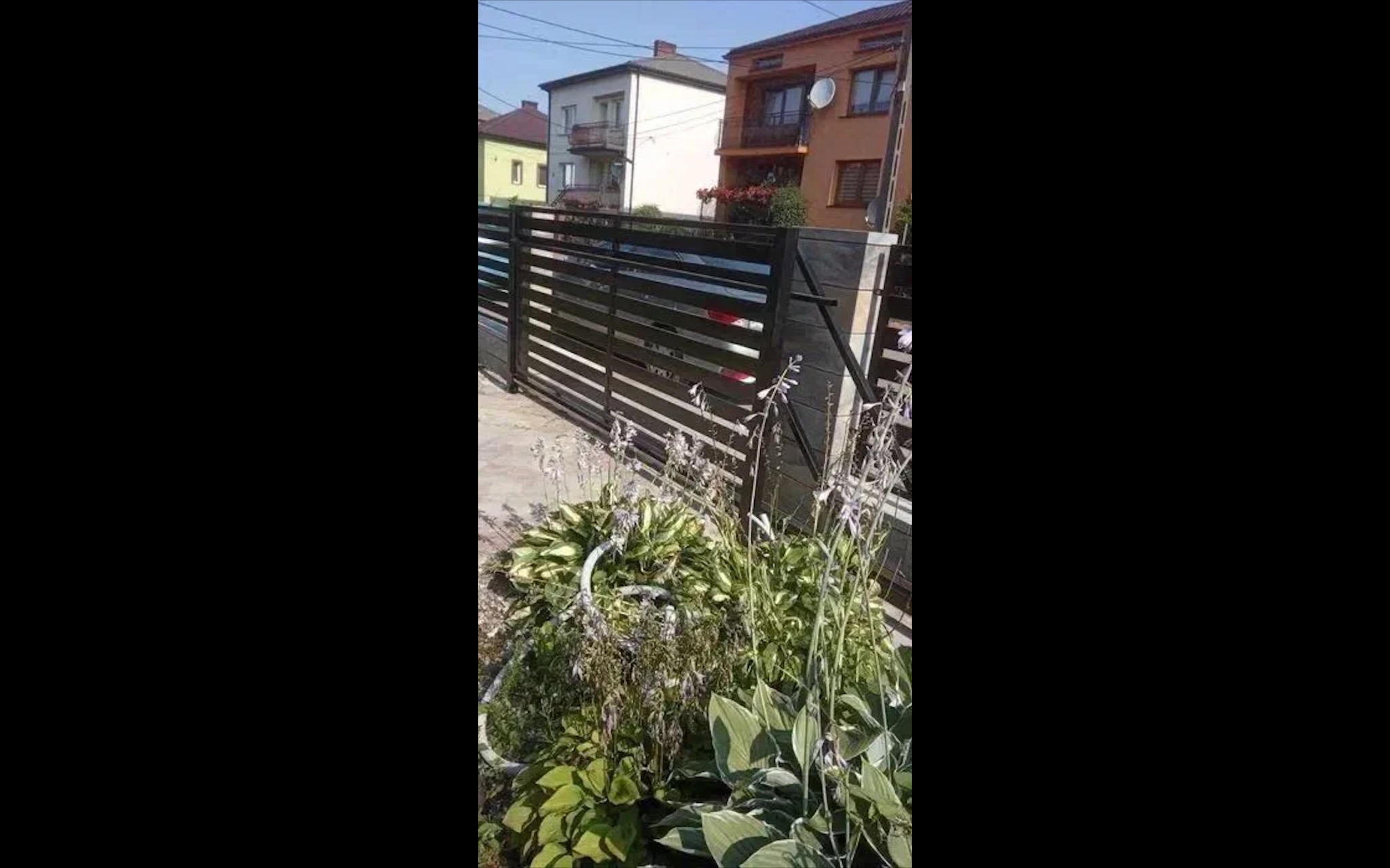 Nowoczesna, pozioma brama wjazdowa z metalu, z widocznymi przęsłami i betonowymi słupkami. Na pierwszym planie roślinność ogrodowa, w tle budynki mieszkalne.