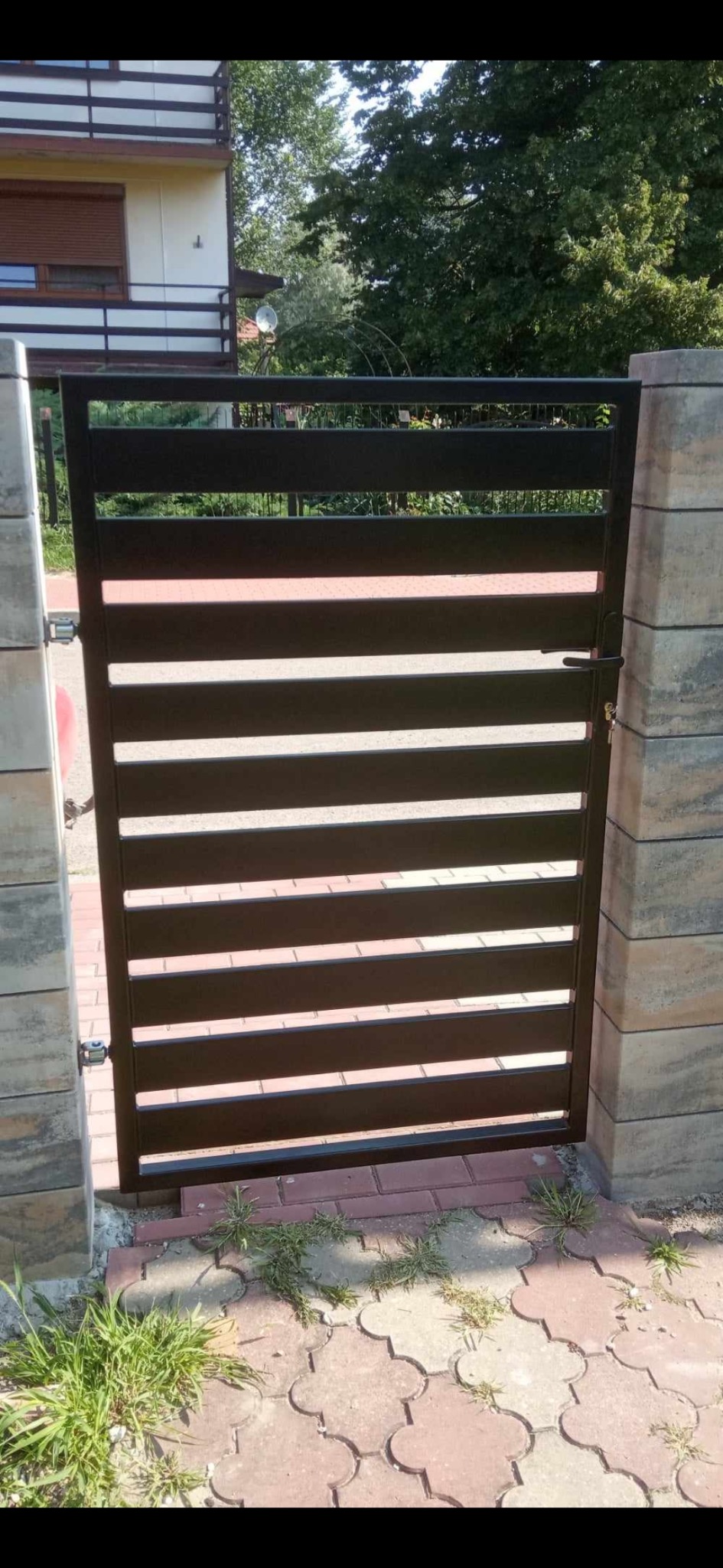 Nowoczesna, brązowa furtka z poziomych profili, zamontowana między kamiennymi słupkami. Widoczne detale zamka i zawiasów. Tło stanowi dom i zieleń.