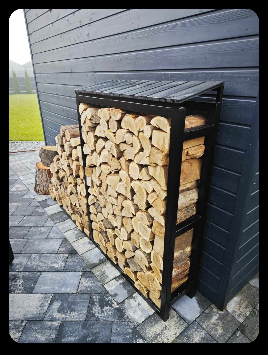 Metalowy regał na drewno opałowe z daszkiem, wypełniony pociętymi kawałkami drewna, stojący na szarym bruku przy ciemnej, drewnianej ścianie budynku.