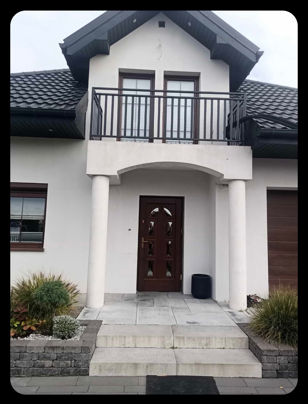 Dwukondygnacyjny dom z balkonem z czarną balustradą, kolumnami przy wejściu i ciemnobrązowymi drzwiami. Widoczne schody wejściowe i roślinność.