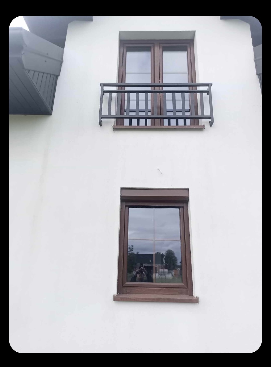 Elewacja budynku z oknem i balkonem zabezpieczonym ciemną, metalową balustradą. W oknie odbija się osoba robiąca zdjęcie.
